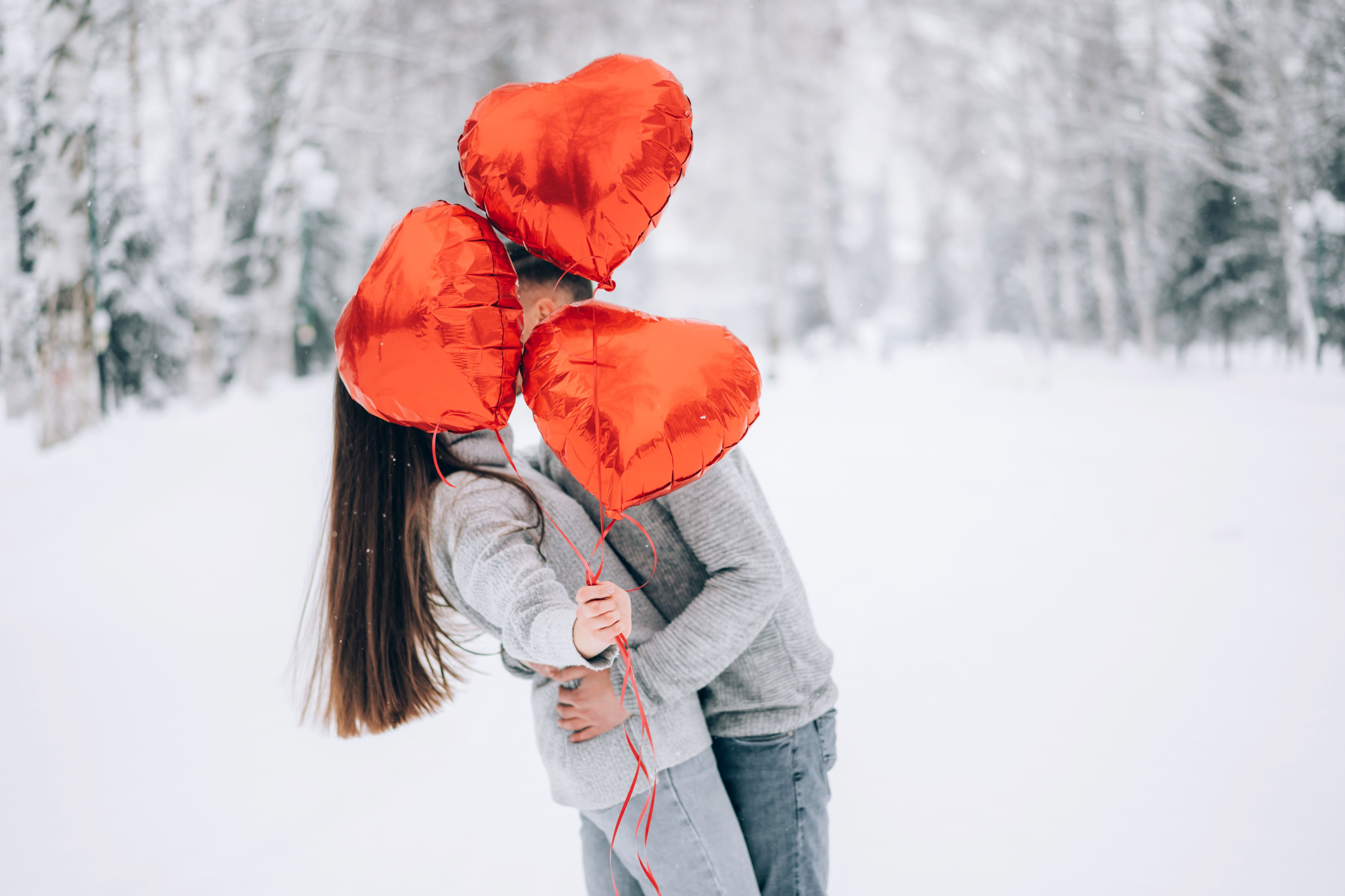 Mies ja nainen suutelevat sydämen muotoisten ilmapallojen takana talvisessa maisemassa.