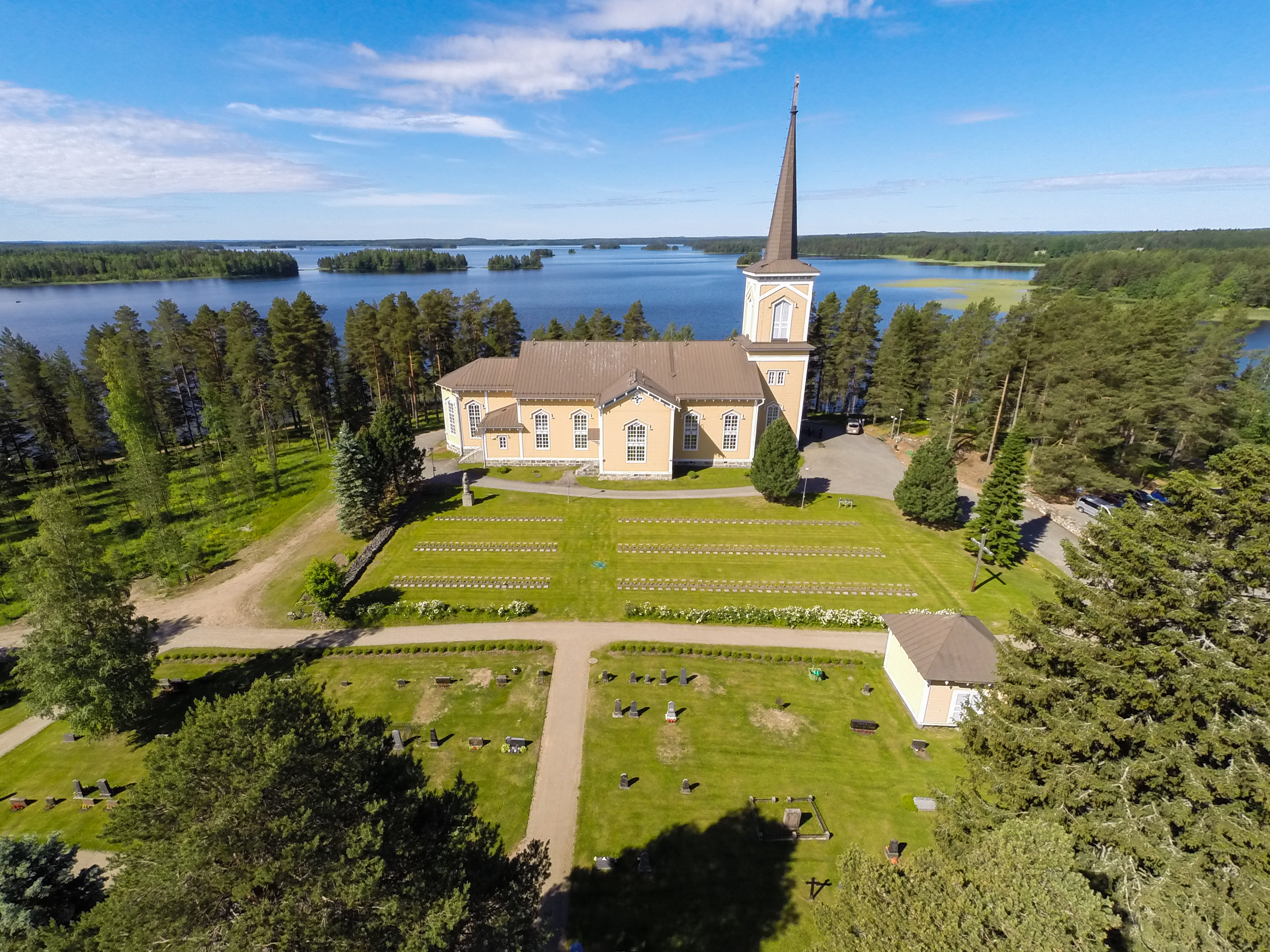 Pielaveden kirkko ilmasta, hautausmaa, kirkko ja järvi takana. Rannassa suuria puita, sininen taivas ja kes...