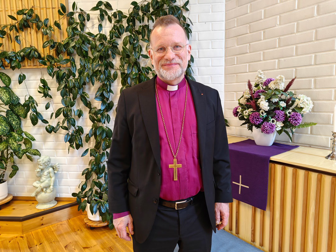 Piispa Jari Jolkkonen alttarilla, katsoo kameraan ja hymyilee.