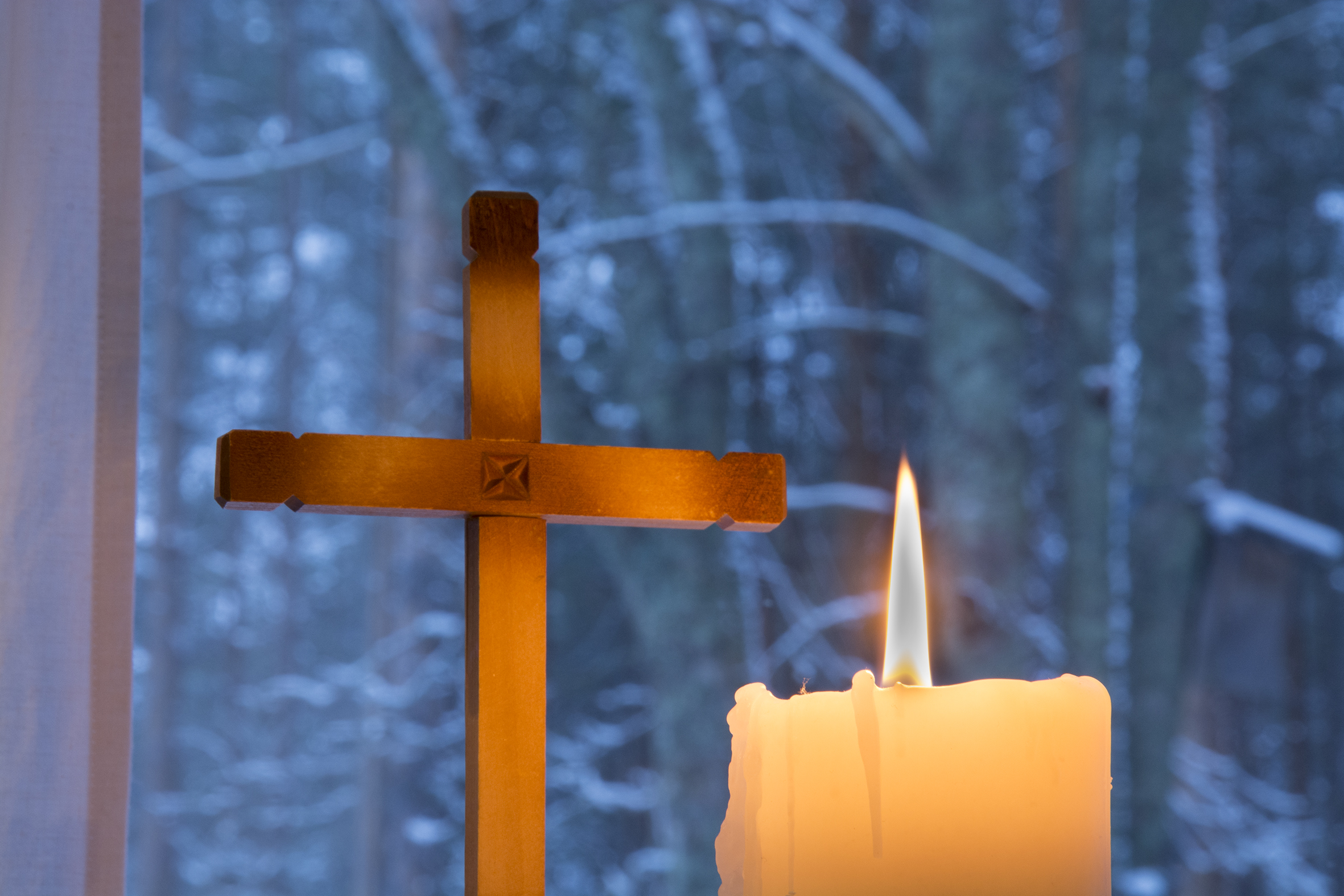 Ntualalla palava valkoinen kynttilä, takana puinen risti. Näkymä ikkunasta lumiseen metsään.